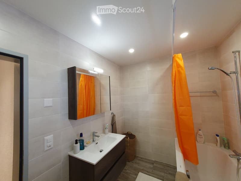 Modern bathroom with a white tiled wall, a dark wooden vanity with a sink, a mirror, a bathtub with an orange shower curtain, and various toiletries on the sink counter.
