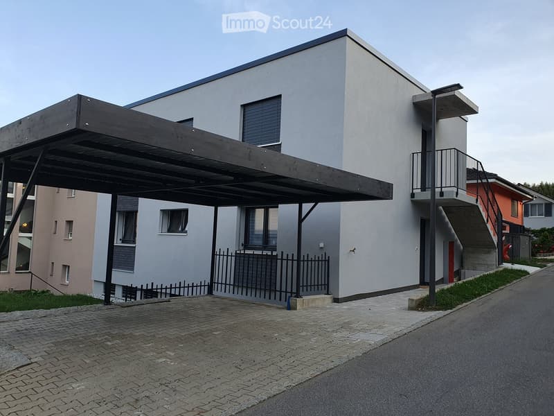 Modern two-story house with a flat roof, large windows, and a covered parking area. The house features a sleek design with a combination of white and grey exterior walls.