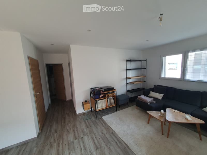 Modern living room with light wooden flooring, white walls, and large windows. Features a dark grey sectional sofa, a small coffee table, a wooden shelf with books and decorative items, and a sleek black shelving unit.