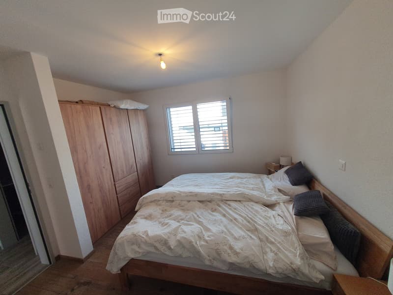 Cozy bedroom with a wooden bed, white bedding, pillows, a nightstand, and a wardrobe. The room features a window with blinds and a slanted ceiling.