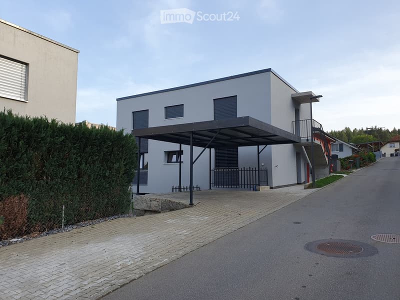 Modern two-story house with a flat roof, grey exterior, and a covered parking area. The house features large windows and a small balcony on the upper level.