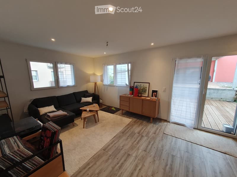 Modern living room with a black sectional sofa, wooden coffee table, side table, floor lamp, and a wooden cabinet with picture frames on top. The room features large windows and a sliding glass door leading to a balcony.