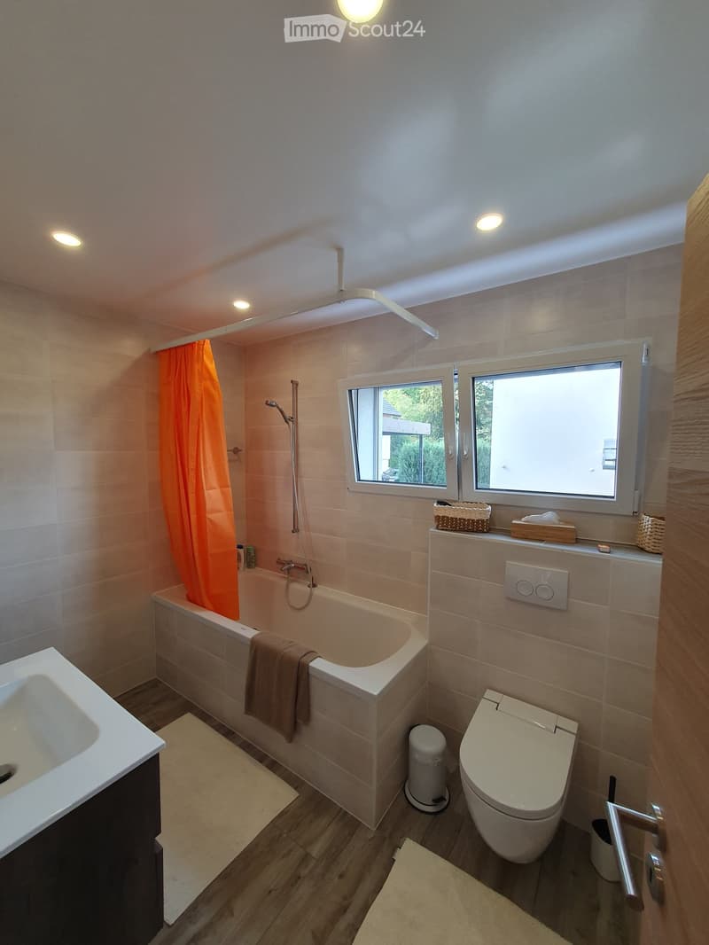 Modern bathroom with a bathtub, toilet, sink, shower curtain, and a small window with a view of greenery outside.