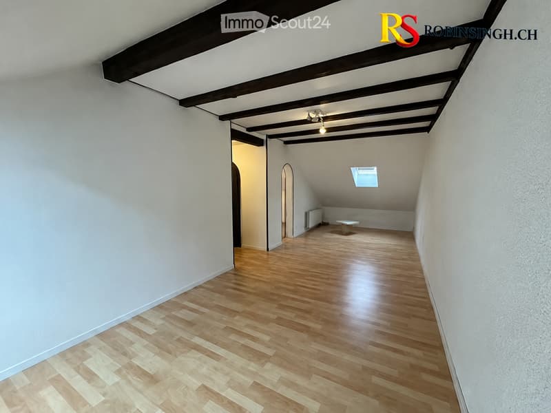 Empty attic room with wooden flooring, white walls, exposed wooden beams on the ceiling, and a small skylight window providing natural light.