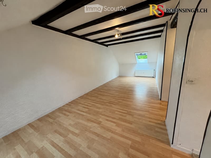 Empty room with wooden flooring, white walls, and exposed wooden beams on the ceiling. A small window at the far end provides natural light.