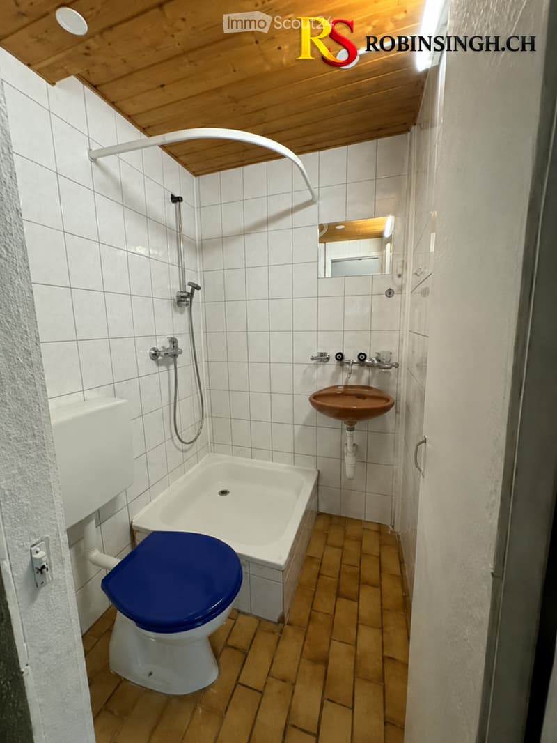 Bathroom with white tiled walls, wooden floor, toilet with blue seat, bathtub, sink, mirror, and showerhead.
