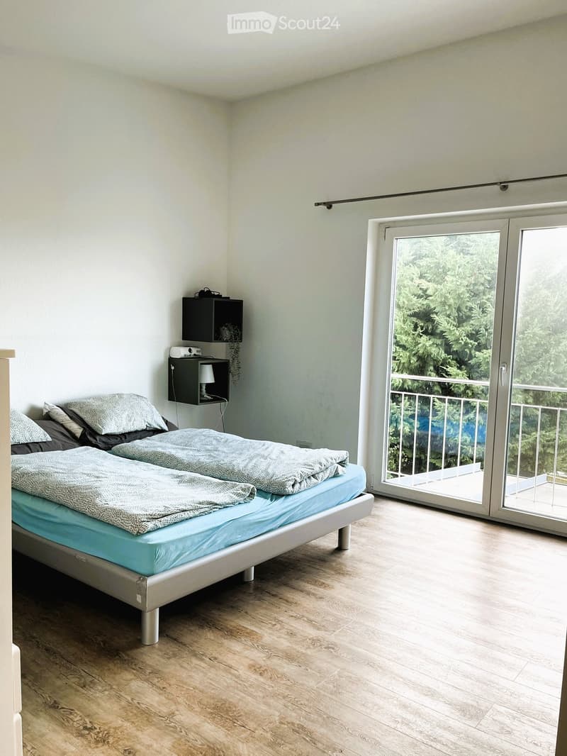 Modern bedroom with a double bed, nightstand, lamp, and a sliding glass door leading to a balcony with a view of greenery.