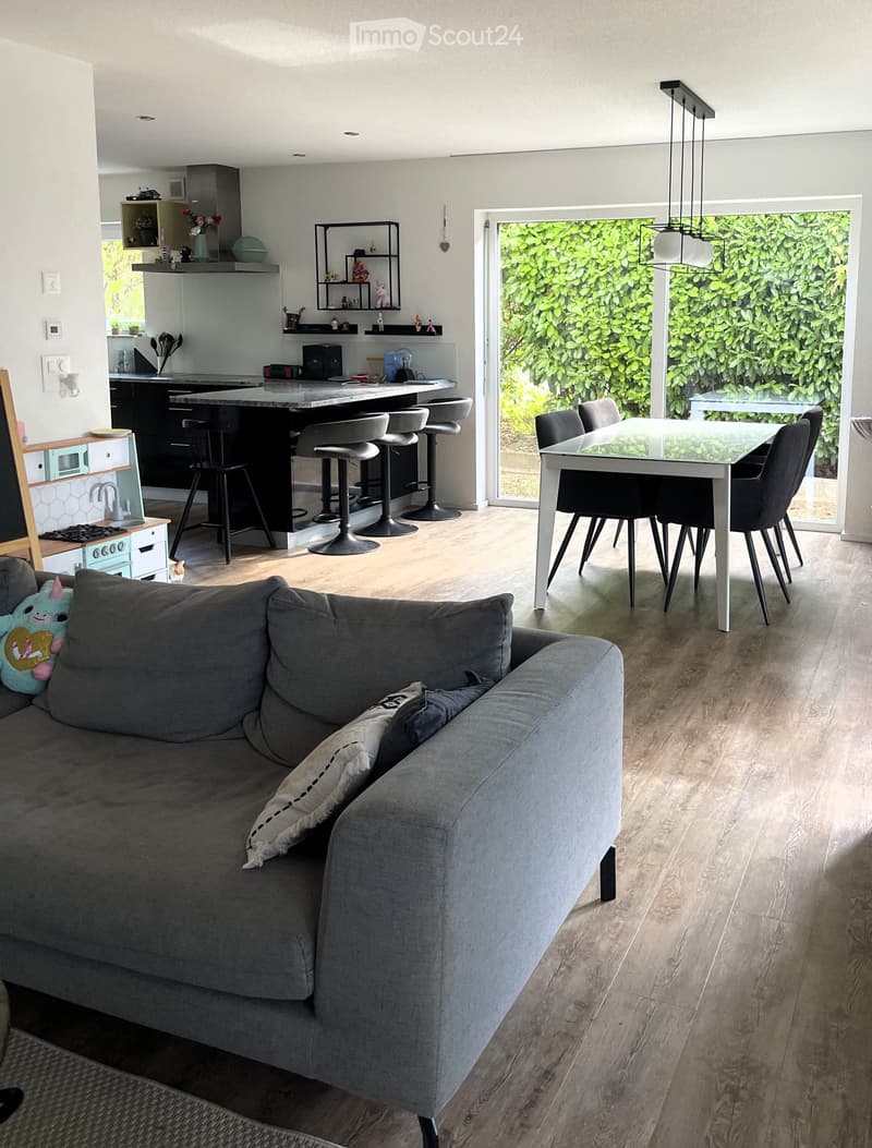 Open-plan living and dining area with a grey sofa, a dining table with chairs, a kitchen island with stools, and a modern kitchen setup.