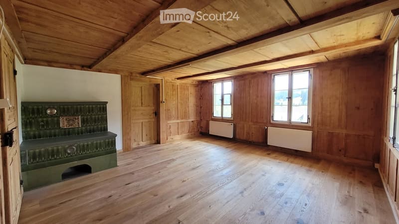 Empty room with wooden walls, wooden ceiling beams, wooden floor, and a green antique bench against one wall. Windows and radiators are visible.