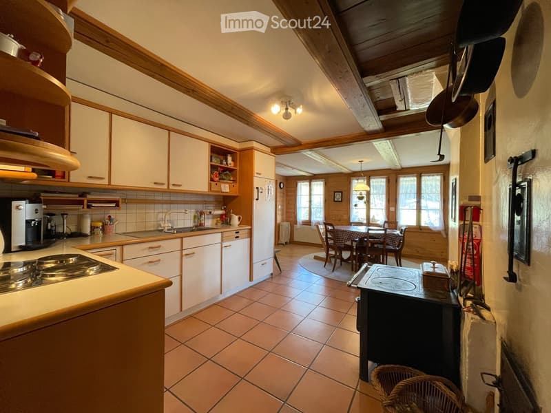 The image shows a cozy, well-lit kitchen with white cabinets, a tiled floor, and a dining area. The kitchen features a sink, stove, coffee maker, and various utensils. A dining table with chairs is visible in the adjacent space, along with a wicker basket