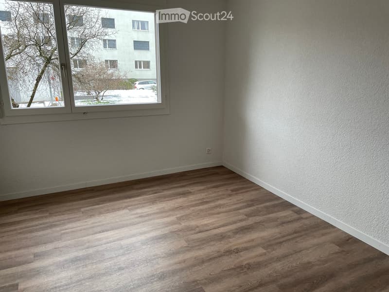 Empty room with wooden flooring and white walls, featuring a large window with a view of the outside.