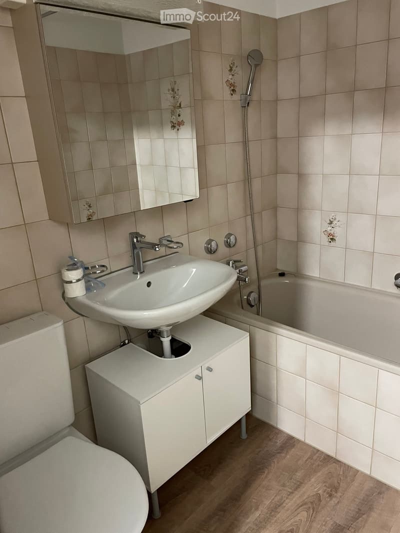Modern bathroom with a white sink, chrome faucet, toilet, bathtub, and a mirrored cabinet above the sink. The walls are tiled with floral designs, and the floor is wooden.