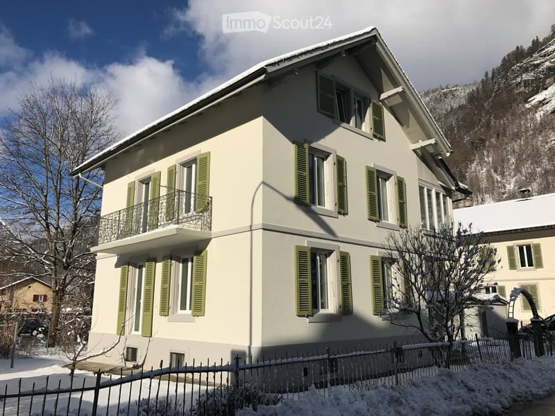 Two-story house with light green shutters, white walls, and a balcony on the second floor. Snow covers the ground and roof.