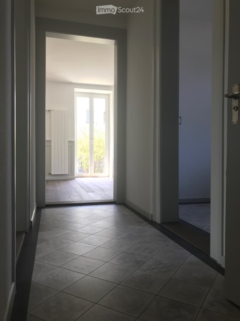 Empty hallway with tiled floor, leading to an open doorway that reveals a bright, sunlit room beyond.