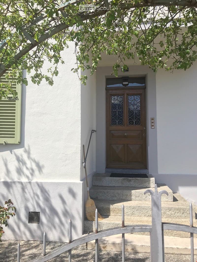 Entrance to a house with a wooden double door featuring decorative glass panels, flanked by white walls and a small staircase leading up to it. A broom leans against the wall beside the steps, and a metal gate is visible in the foreground.