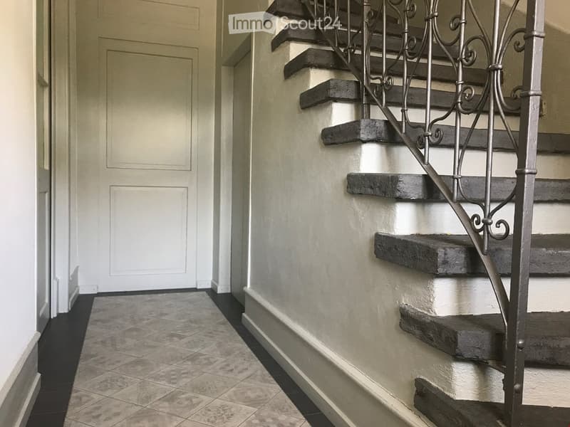 A hallway with a staircase featuring dark wooden steps and a decorative metal railing, light-colored walls, and tiled flooring.