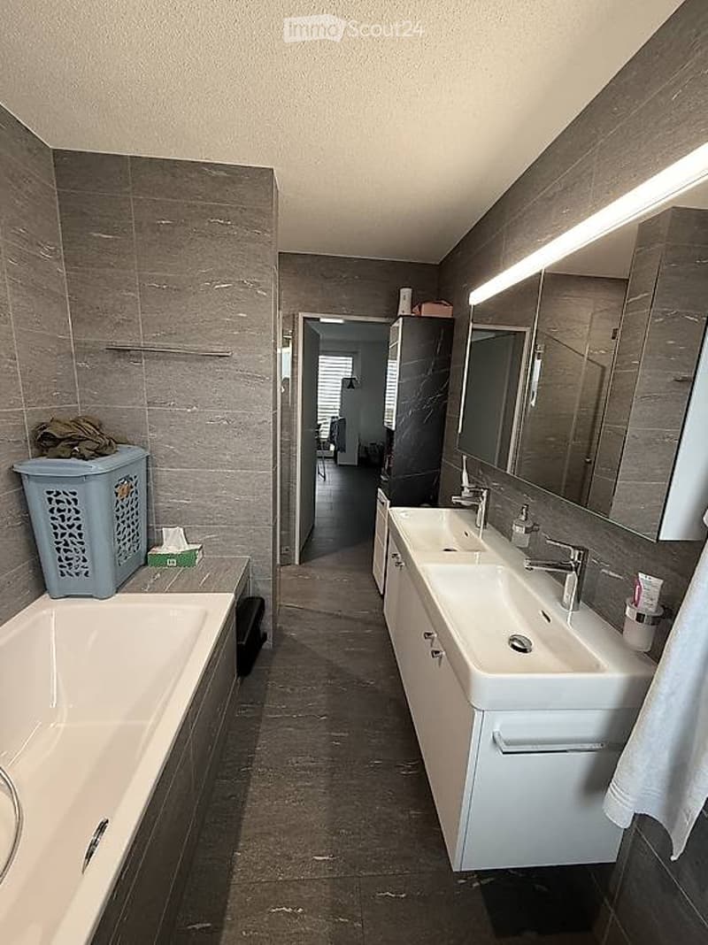 Modern bathroom with a bathtub, double sink, large mirror, and built-in cabinets. Laundry basket and cleaning supplies are visible.