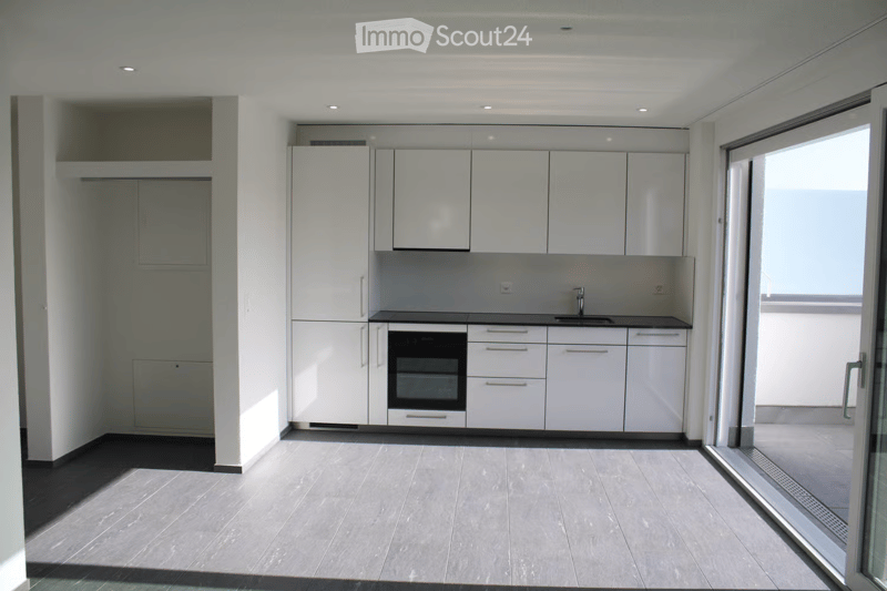 Modern, minimalist kitchen with white cabinets, a built-in oven, and a sink, featuring a large window and a sliding glass door leading to a balcony.