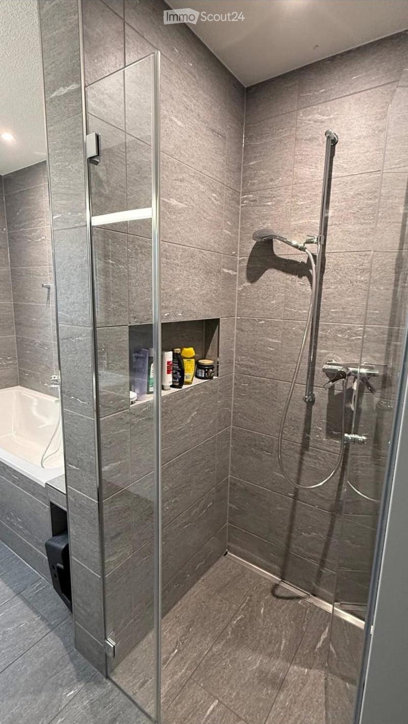 Modern bathroom with grey tiled walls and floor, featuring a bathtub, glass shower enclosure, and a shower with a handheld sprayer.