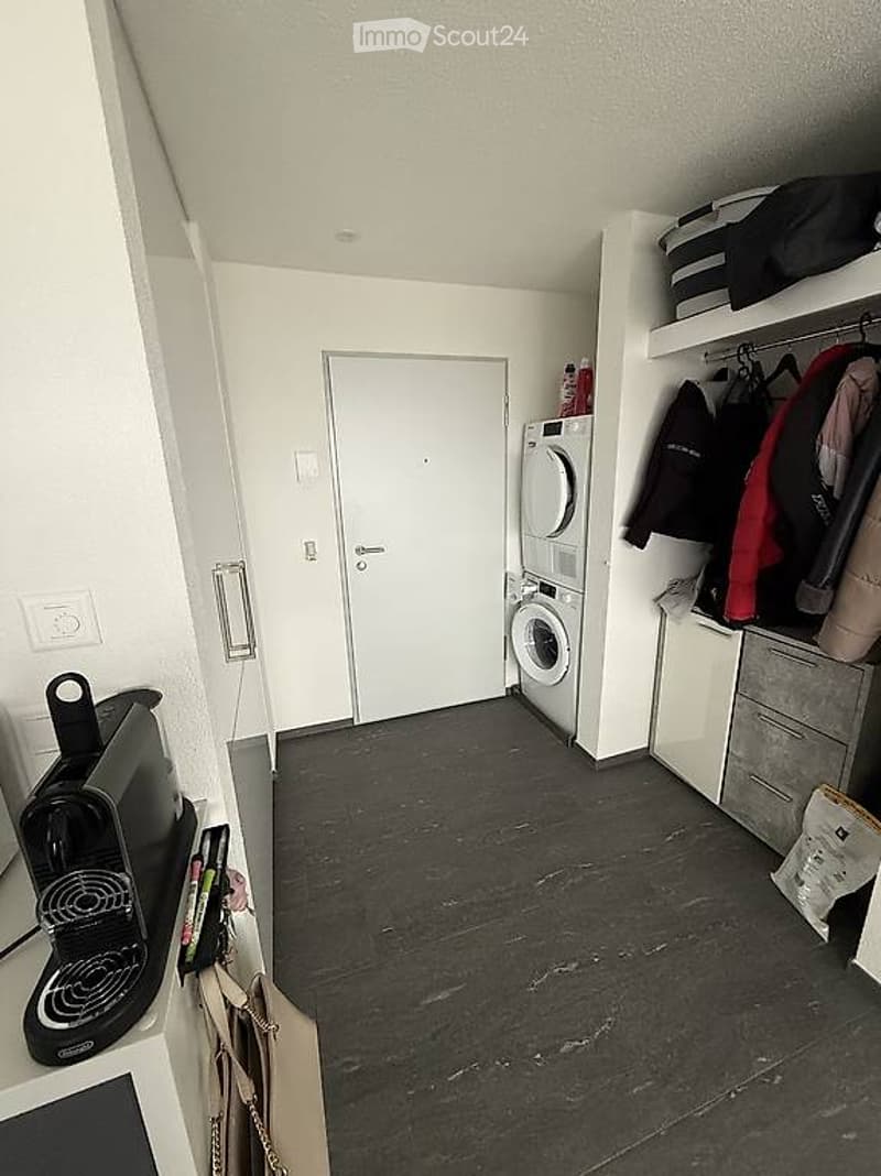 A small, modern hallway with a dark grey tiled floor, white walls, and a closed white door. A black coffee maker sits on a counter to the left, and a washing machine is positioned against the wall to the right. A built-in wardrobe with hanging coats and a