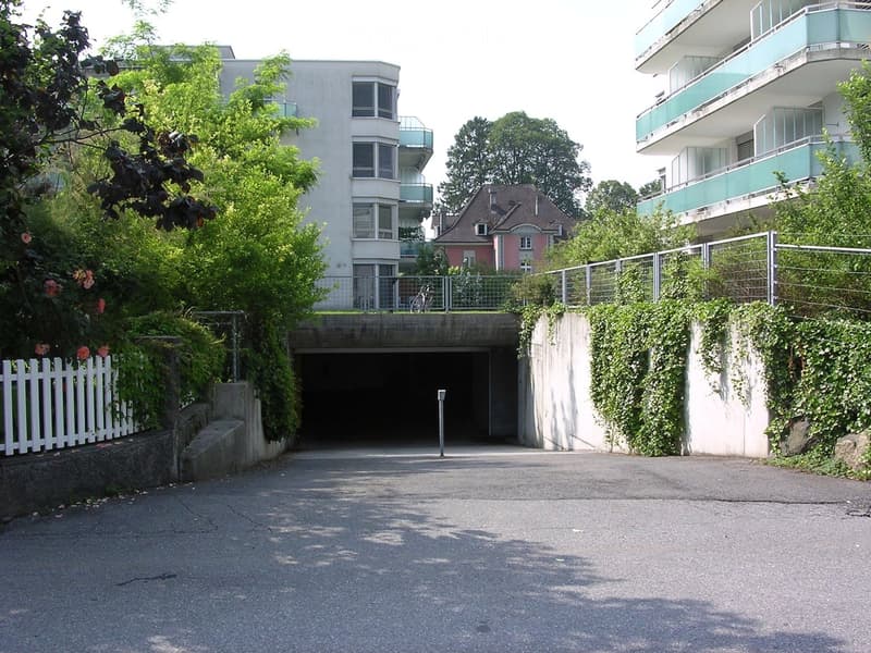 Modern multi-story residential building with balconies, surrounded by greenery and a gated entrance leading to a parking area.