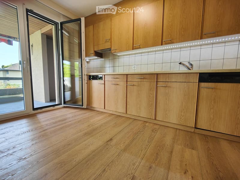 Modern kitchen with light wooden cabinets, a sleek countertop, and a built-in dishwasher. The space features a polished wooden floor and a large sliding glass door leading to an outdoor area.
