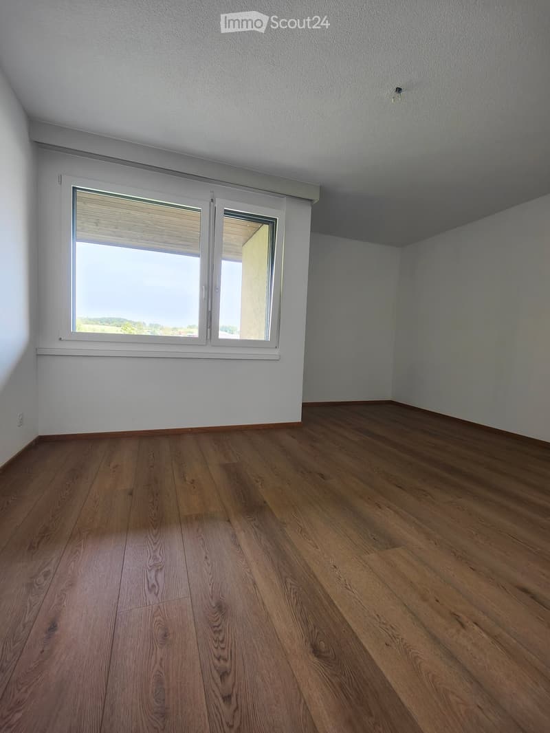 Empty room with wooden flooring and white walls, featuring a large window with blinds and a view of the outdoors.