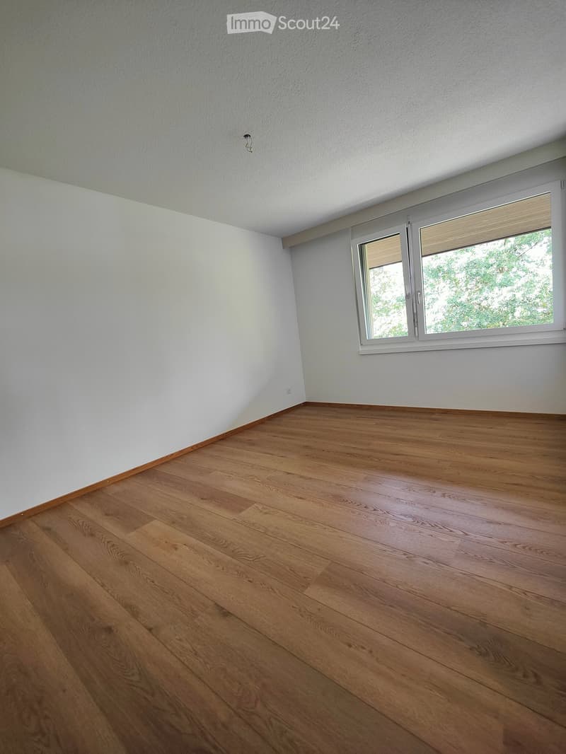 Empty room with light-colored walls and wooden flooring, featuring a large window with blinds.