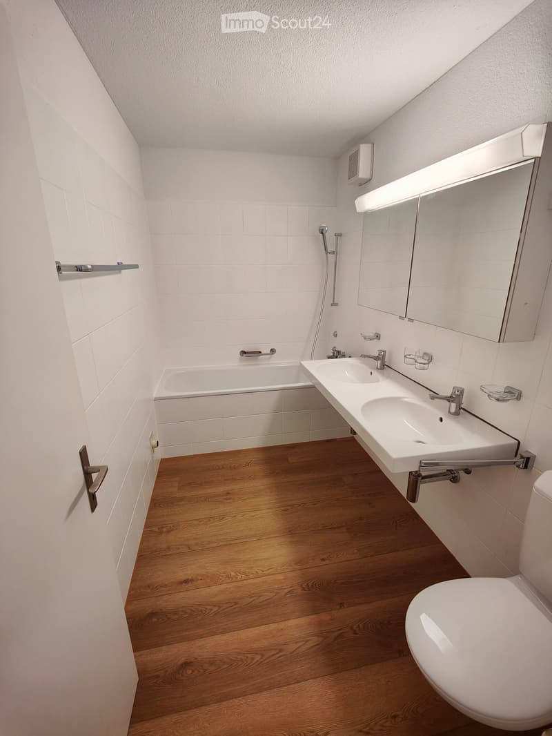 Modern bathroom with white tiles, wooden floor, bathtub, double sink, toilet, and large mirror with built-in lighting.