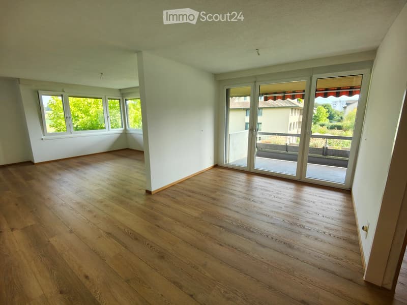 Empty room with wooden flooring, white walls, large windows, and sliding glass doors leading to a balcony.