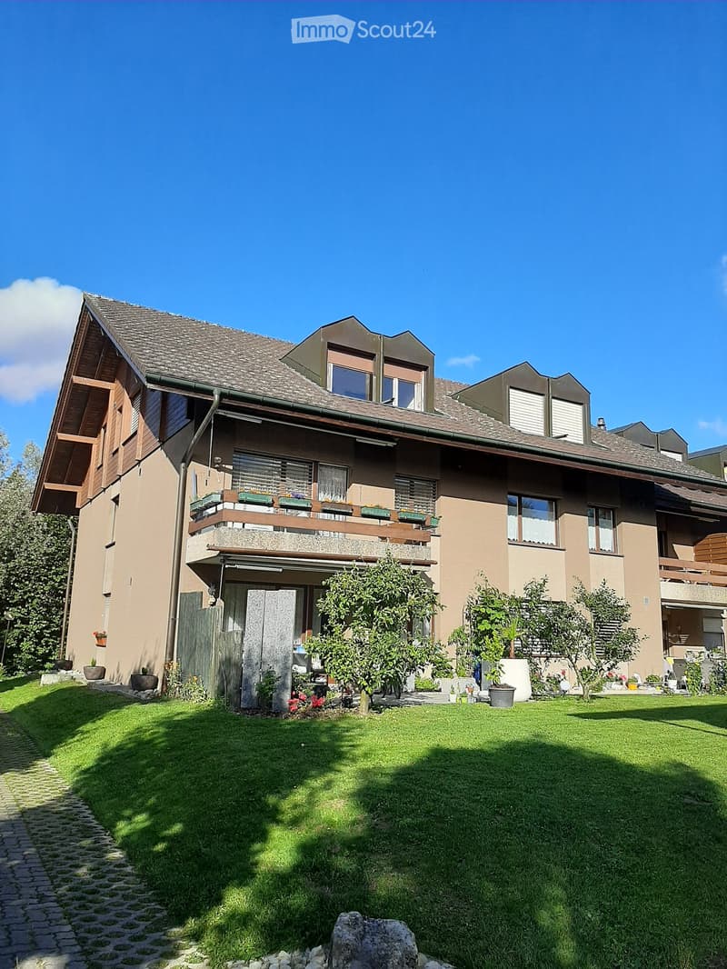 Two-story house with a balcony, multiple windows, and a well-maintained lawn with trees and shrubs.