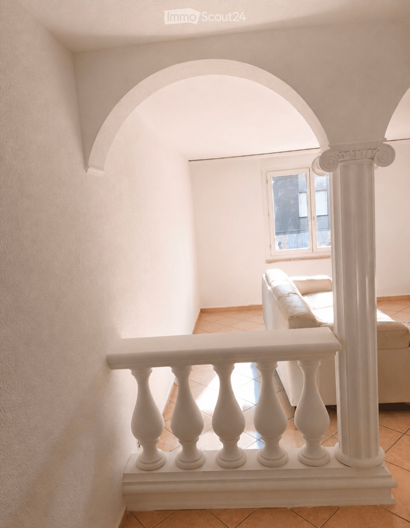 A white balustrade with decorative columns leads to a room with a beige sofa and a window offering a view of the outside.