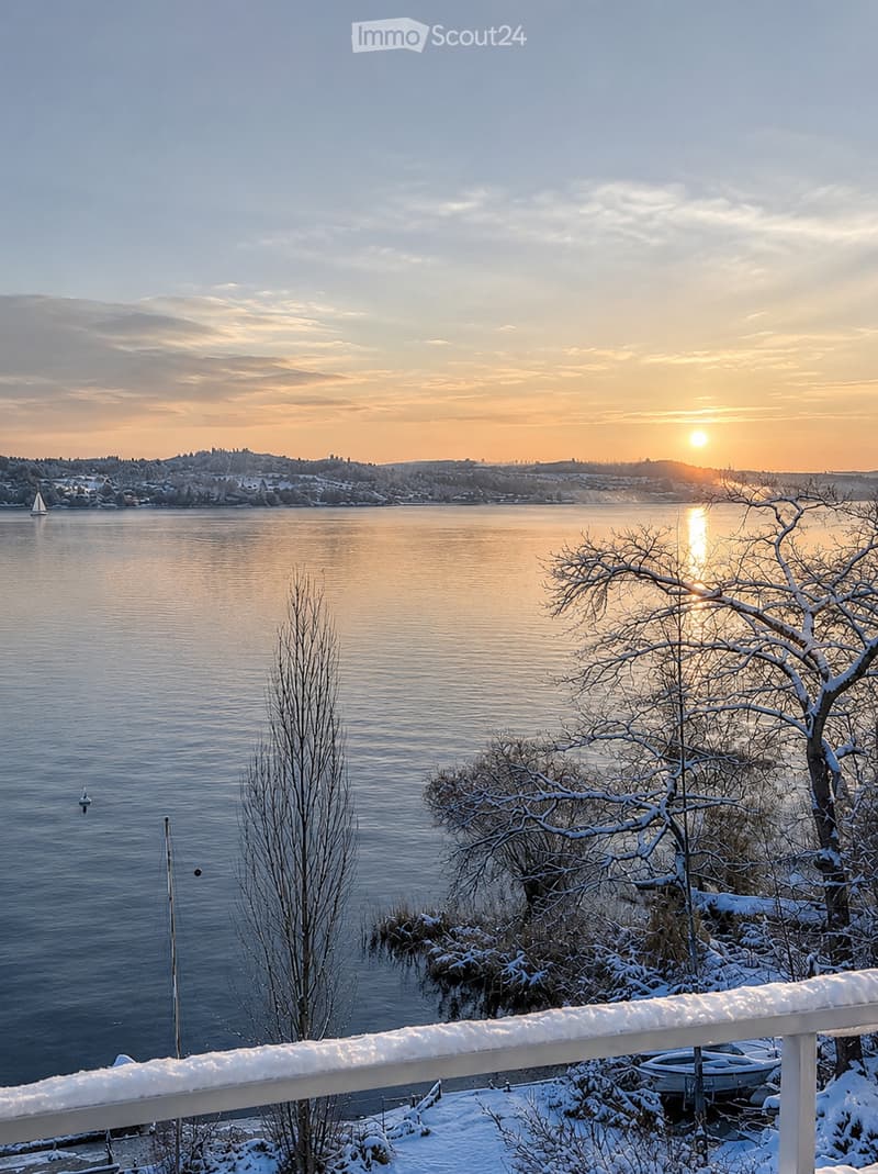 Snow-covered balcony overlooking a serene lake at sunset, with distant cityscape and sailboat visible.