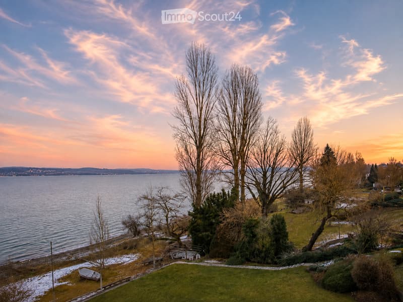 View of a waterfront property with a grassy lawn, bare trees, a small dock, and a calm body of water under a sunset sky.