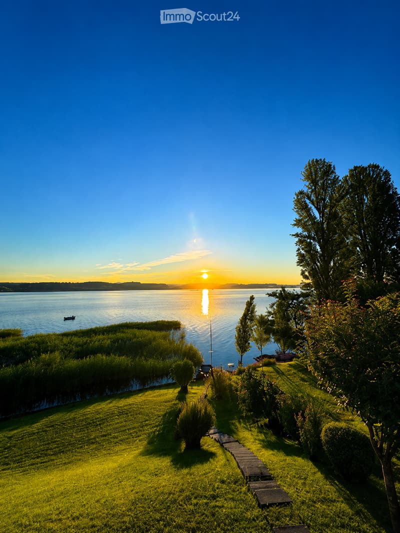 A well-maintained garden with a stone pathway leading to a waterfront view, featuring lush greenery, trees, and a serene lake with a sunset reflection.