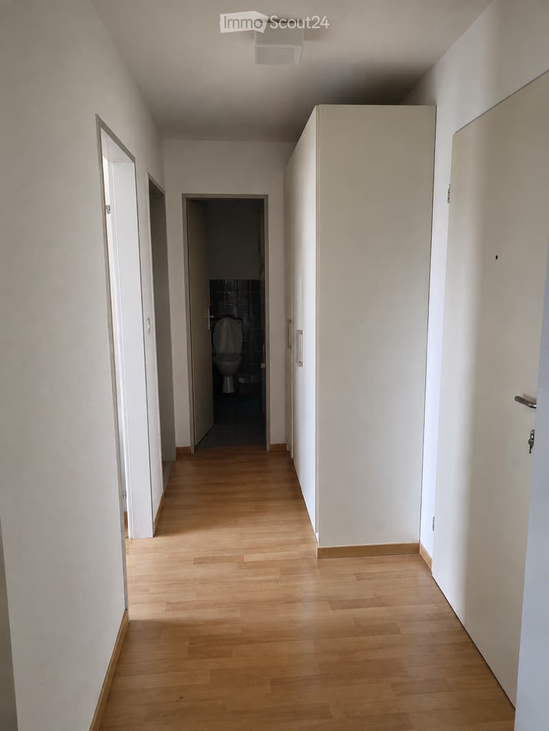 Empty hallway with light wooden flooring, white walls, and a visible bathroom at the end.