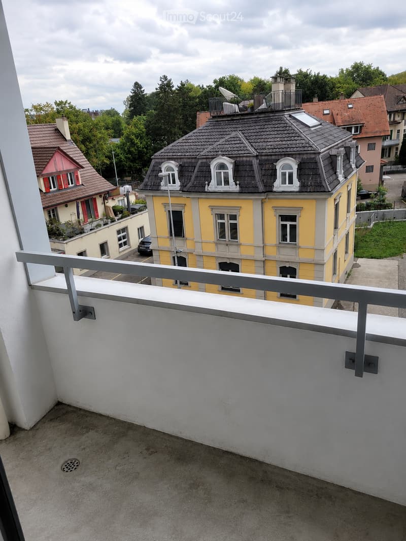 Balcony with metal railing, overlooking a residential area with colorful houses, trees, and a parked car.