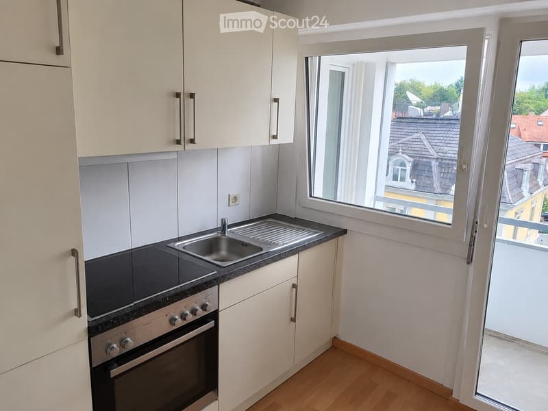 Modern kitchen with light-colored cabinets, a stainless steel sink, and an electric stovetop oven. Large window offers a view of the city.