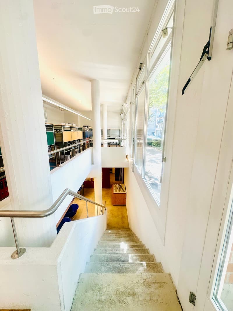 Modern interior staircase with white walls, large windows, metal handrail, and visible furniture in the background including chairs and a table.