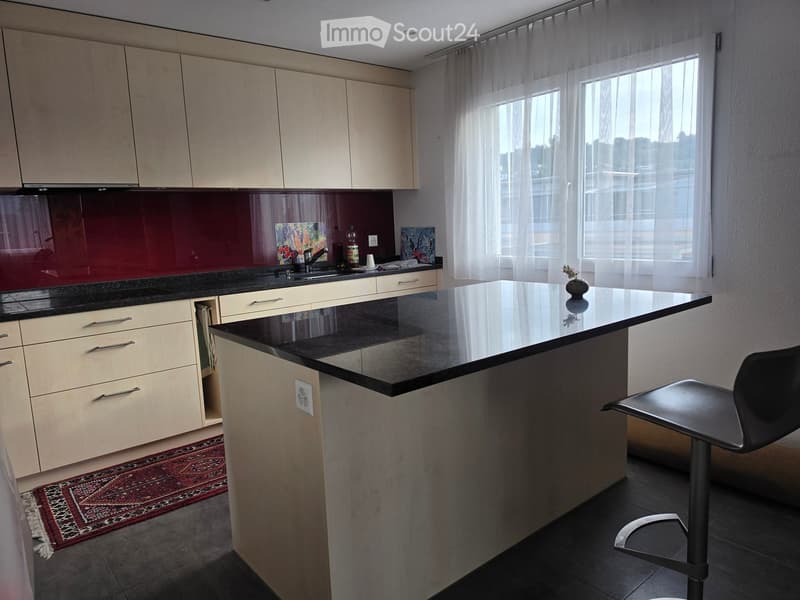 Modern kitchen with light wood cabinets, black granite countertop, sink, and a small island with a stool. Large windows with sheer curtains provide natural light.