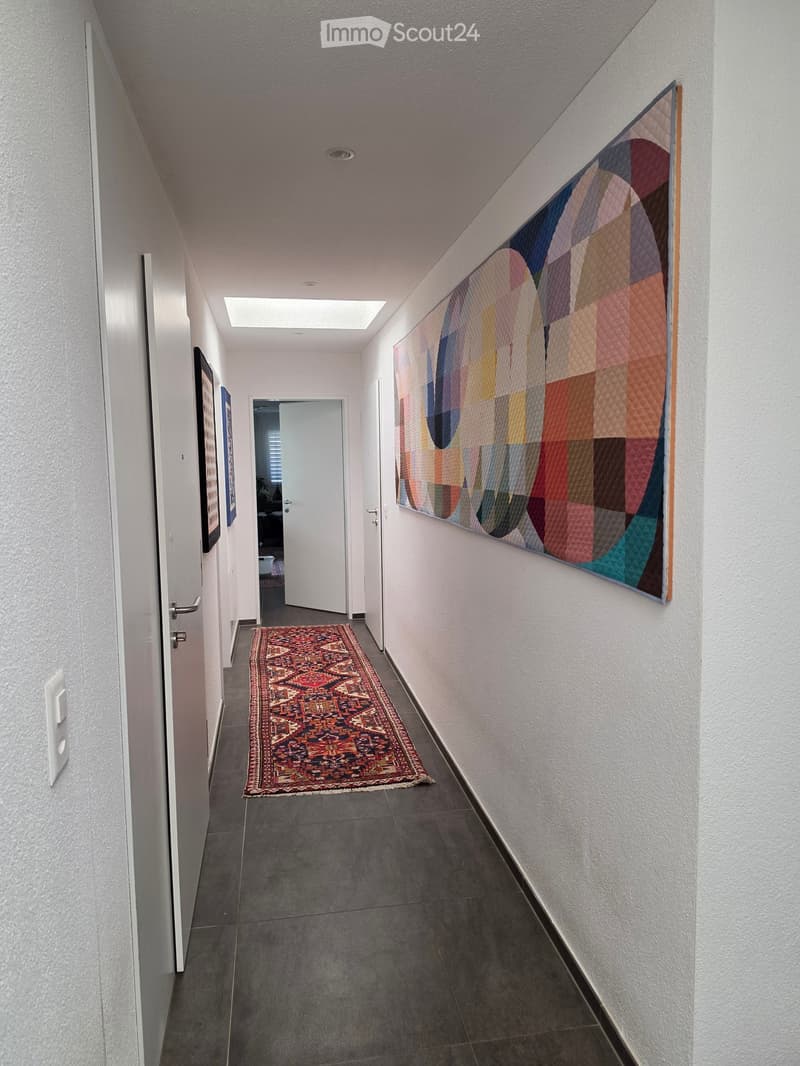 Clean, modern hallway with grey tiled floor, white walls, and a colorful abstract quilt hanging on one wall. A patterned rug lies in the center, and several doors are visible along the corridor.