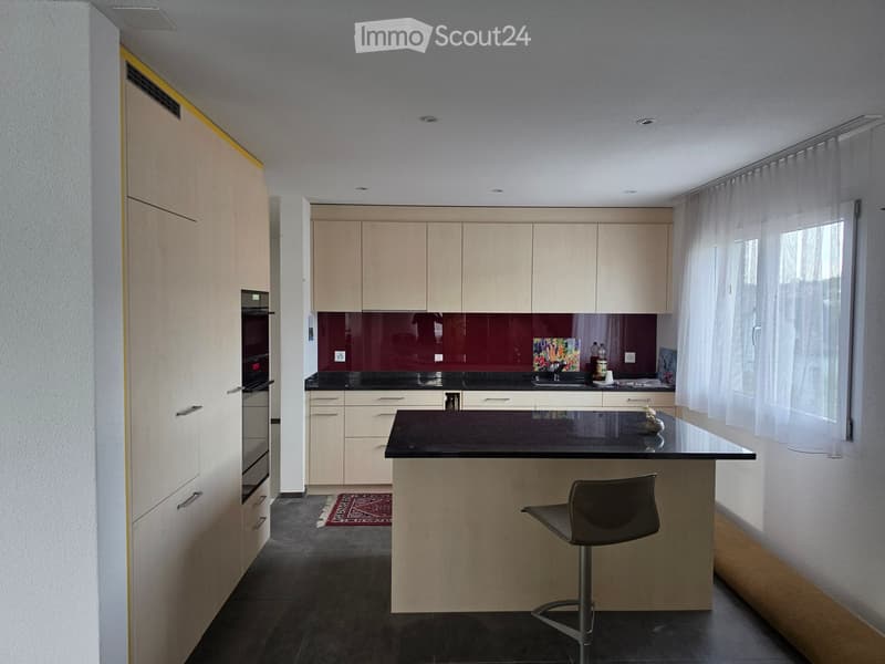 Modern kitchen with light wood cabinets, black countertops, built-in oven, and a central island with a bar stool. Features a dark backsplash and a small decorative rug on the floor.