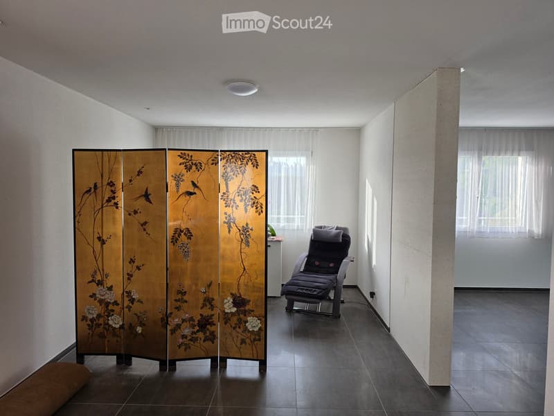 Minimalist room with dark tiled floor, white walls, large windows with sheer curtains, a decorative screen with floral design, and a single massage chair.