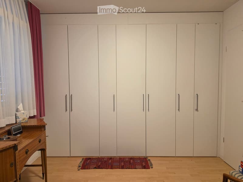 A furnished room with a large white wardrobe, a wooden desk with a small screen, a patterned rug, and a curtain on the window.