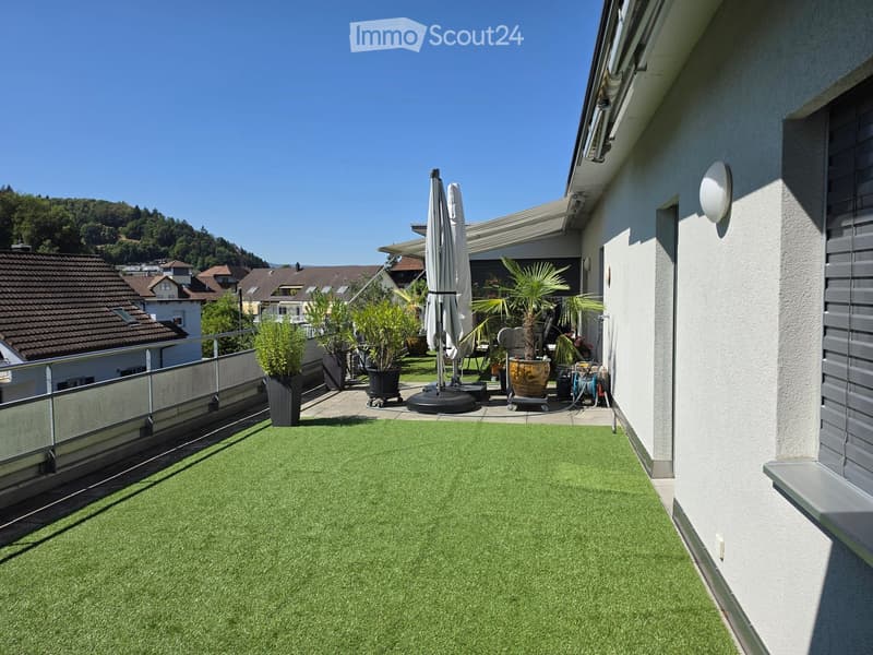 Balcony with artificial grass, potted plants, and an umbrella, overlooking a residential area with houses and greenery.