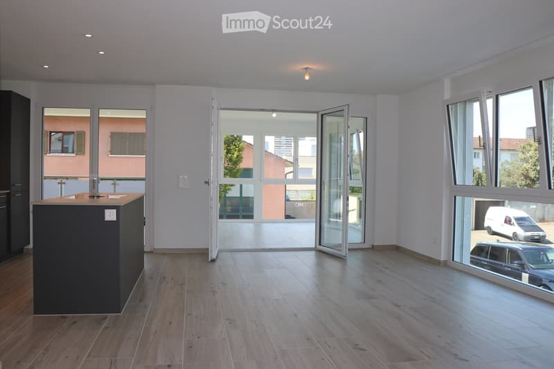 Empty room with wooden flooring, white walls, and large windows with sliding glass doors leading to a balcony. A kitchen island is visible on the left side.