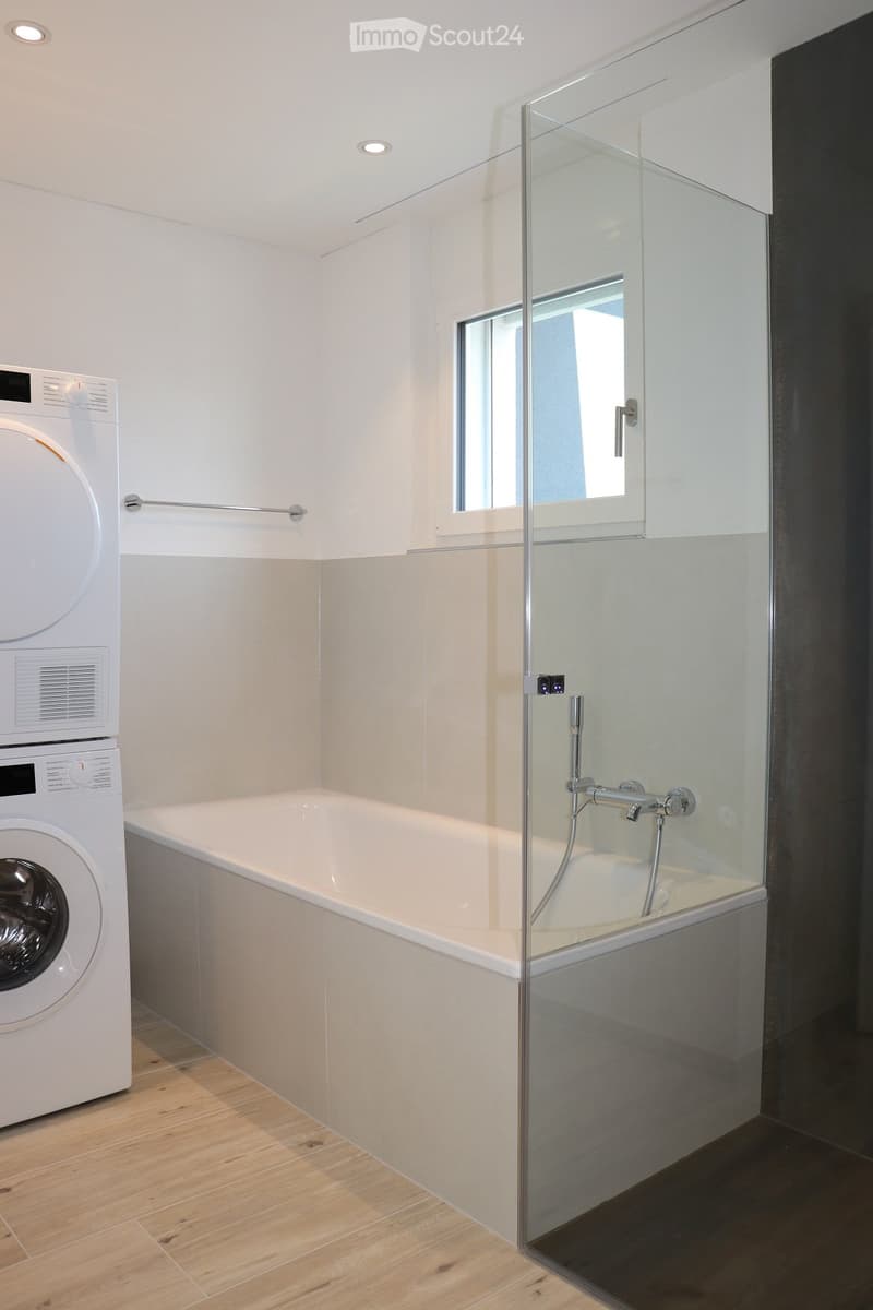 Modern bathroom with a bathtub, glass-enclosed shower, and a washing machine. Features light-colored tiles and wooden flooring.