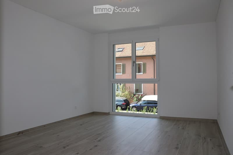 Empty room with wooden flooring and large window showing a view of a neighboring building and parked cars.
