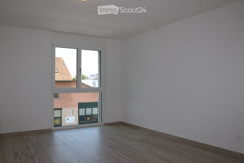 Empty room with light-colored walls and wooden flooring, featuring a large window with a view of a neighboring building.