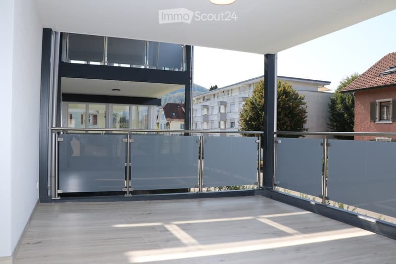 Modern balcony with wooden flooring, glass railing, and a view of surrounding buildings and greenery.